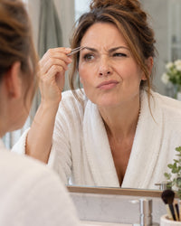 Woman squinting at stray brow hairs in a mirror, tweezers in hand