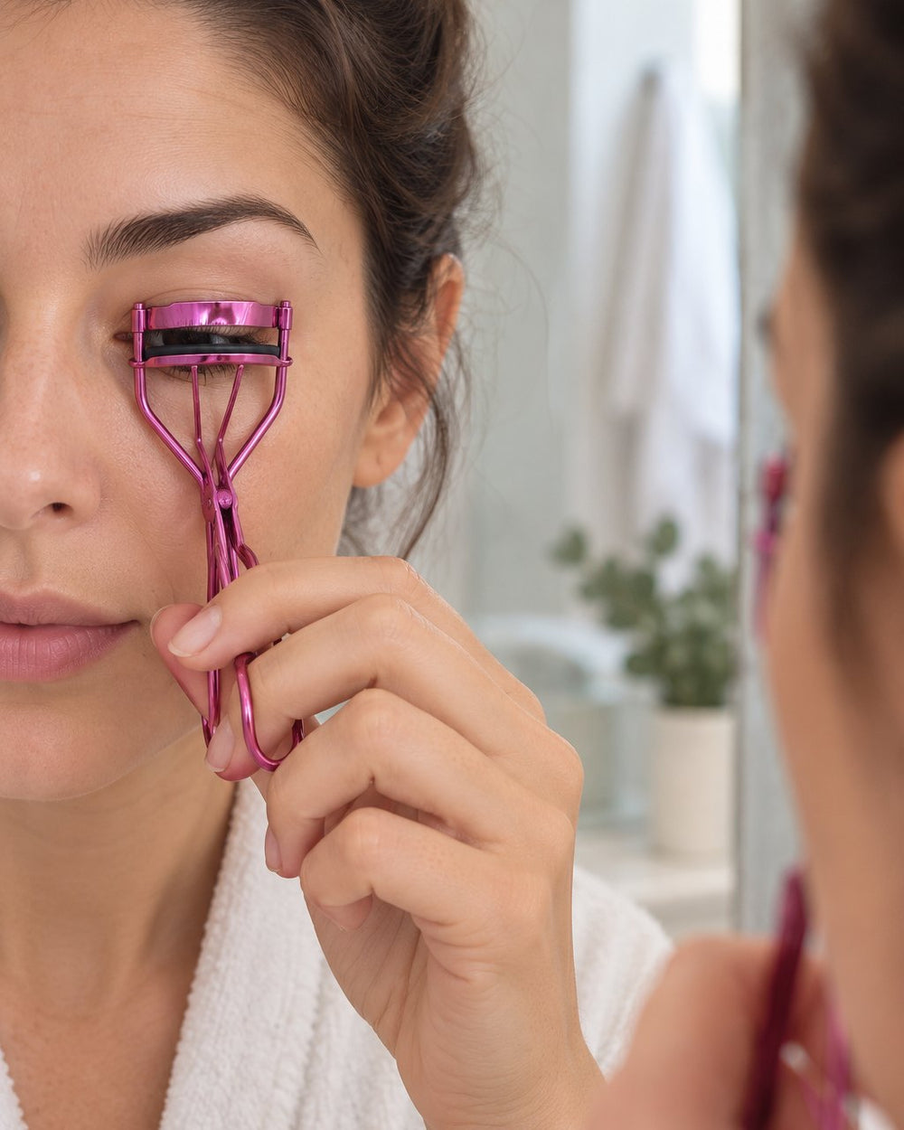 Woman curling her upper lashes with the pink metal curler in a mirror