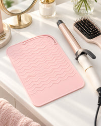 Pink heat mat on a vanity beside a curling wand and a hairbrush