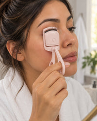 Woman clamping the heated curler on her upper lashes in front of a bathroom mirror
