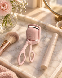 Pink heated lash curler on a vanity tray next to a mascara and a makeup brush