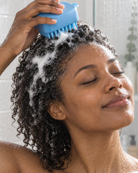 Woman working the blue scalp brush through shampooed hair in the shower